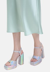 Sandales à plateforme holographiques avec des talons larges, présentant une large sangle avant et une boucle à la cheville, sur une robe en satin menthe.