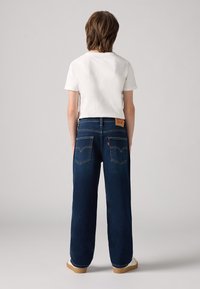 Jeans a gamba larga blu scuro con vita alta, tasche posteriori con cuciture gialle e una patch in pelle con il marchio sulla cintura. T-shirt bianca.