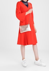 Rød kjole til knæet med klokkeærmer, parret med hvide sneakers. Accessories inkluderer en lille, metallisk guld crossbody taske med en kvast.