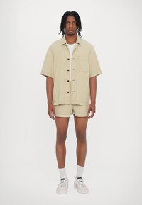 Hellbeige Kurzarm-Hemd mit Knopfleiste und einer Brusttasche, kombiniert mit passenden Shorts. Getragen mit weißen Sneakers und Knöchelsocken.