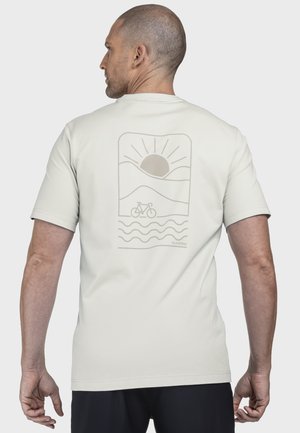 Mann mit einem hellfarbenen T-Shirt, auf dessen Rücken ein Grafikdruck mit Sonne, Hügeln, Wellen und einer Fahrradkontur zu sehen ist.
