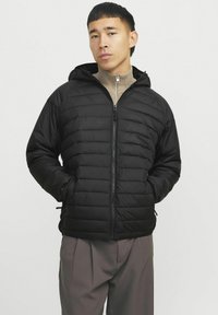 Chaqueta de plumas negra con cremallera frontal, capucha y bolsillos laterales. Presenta un diseño acolchado y una textura suave. Combinada con un suéter beige sin cuello.