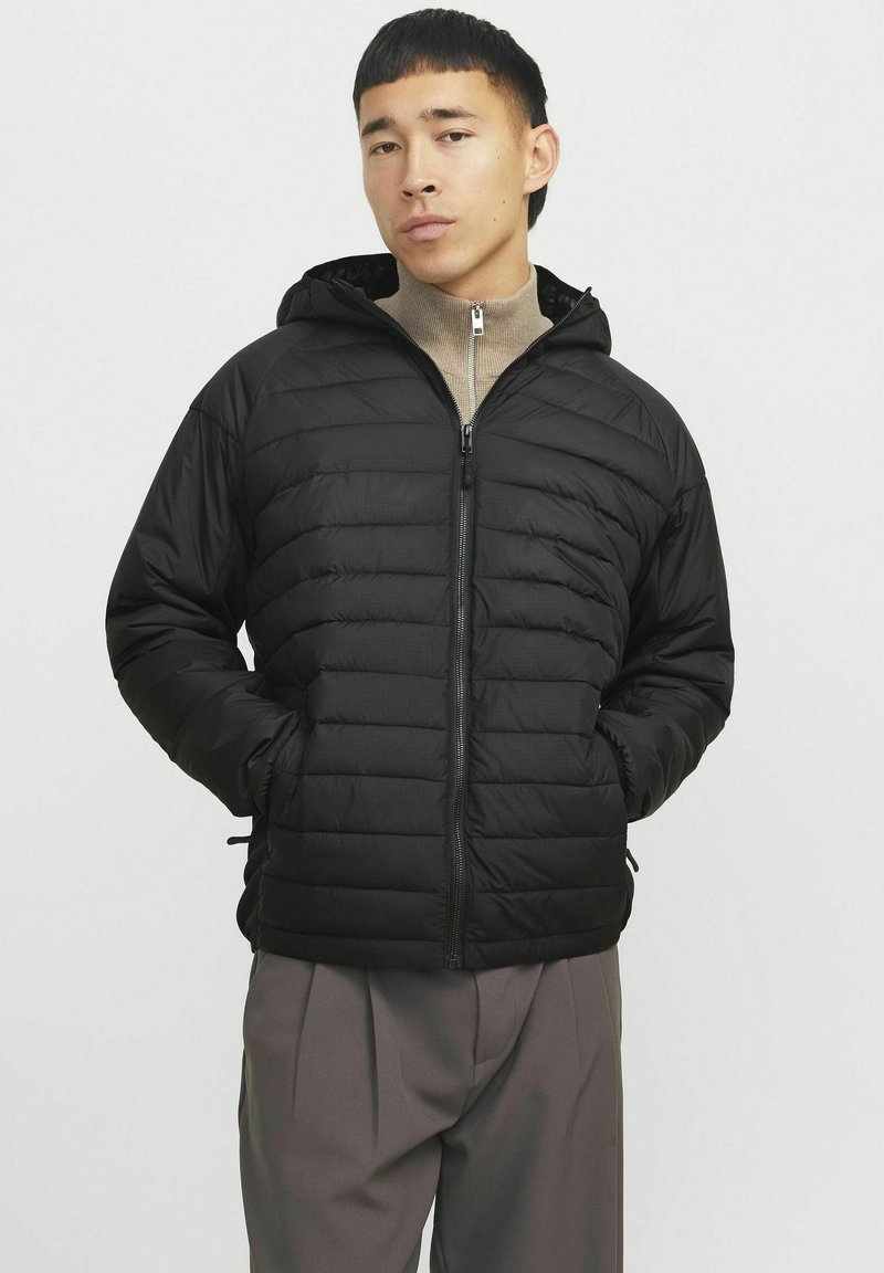 Chaqueta de plumas negra con cremallera frontal, capucha y bolsillos laterales. Presenta un diseño acolchado y una textura suave. Combinada con un suéter beige sin cuello.