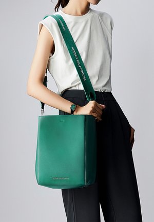 Femme portant un haut blanc sans manches et un pantalon noir, avec un sac à bandoulière en cuir vert à large sangle portant l'inscription « VICTORIA HYDE LONDON ».