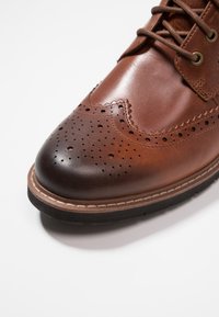 Zapato de cuero marrón con punta redondeada, detalles de brogue y un acabado suave. Presenta una entresuela color tostado y suela de goma negra texturizada.