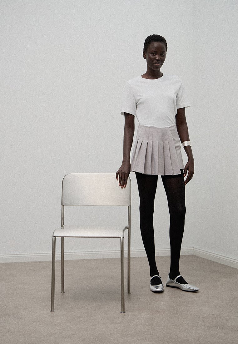 Biała koszulka, jasnoszara plisowana spódnica, czarne rajstopy i srebrne buty. Metalowe krzesło obok modelki w minimalistycznym otoczeniu.