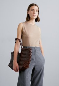 Borsa tote in tessuto marrone con una finitura testurizzata e due manici robusti, indossata sopra una maglietta senza maniche beige e pantaloni grigi su misura.