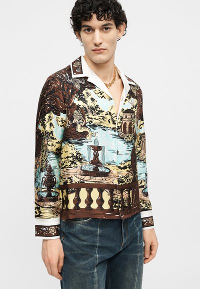Νέος άνδρας που φοράει πουκάμισο με μακριά μανίκια και κουμπιά, διακοσμημένο με πολύχρωμο μοτίβο κήπου και σιντριβανιού, συνδυασμένο με μπλε τζιν και χρυσή αλυσίδα στον λαιμό.
