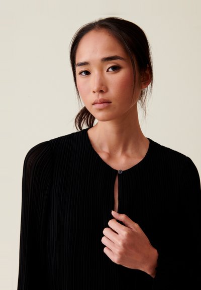 Blouse noire plissée avec un col rond et une fermeture à boutons, dotée de longues manches avec des poignets froncés et une texture lisse.
