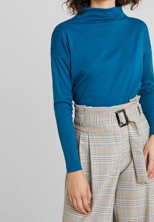Mujer con una blusa azul de manga larga y cuello simulado, metida en pantalones beige de cuadros de cintura alta, con un cinturón negro con hebilla, de pie frente a un fondo gris.