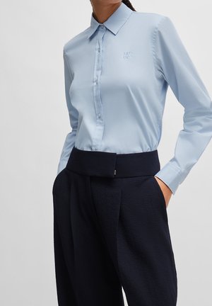 Frau trägt ein hellblaues Hemd mit Knöpfen und besticktem "HUGO"-Logo, kombiniert mit hoch taillierten, weit geschnittenen schwarzen Hosen, die Hände in den Taschen.