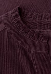 Tkanina sztruksowa w głębokim kolorze śliwkowym ma falbaniasty dekolt. Tekstura jest prążkowana, z widocznymi szwami wzdłuż brzegów.