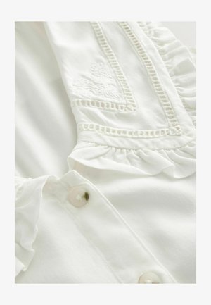 White blouse featuring embroidered detailing, ruffled edges, and button closures. Soft fabric with a smooth texture and delicate accents.