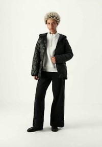 Pessoa em pé a usar uma jaqueta acolchoada preta sobre uma blusa branca com fecho éclair, calças largas pretas e sapatos pretos contra um fundo simples.