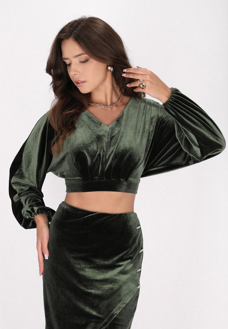 Top corto in velluto verde scuro con maniche ampie e scollatura a V, abbinato a una gonna coordinata con dettagli argento e un design drappeggiato.
