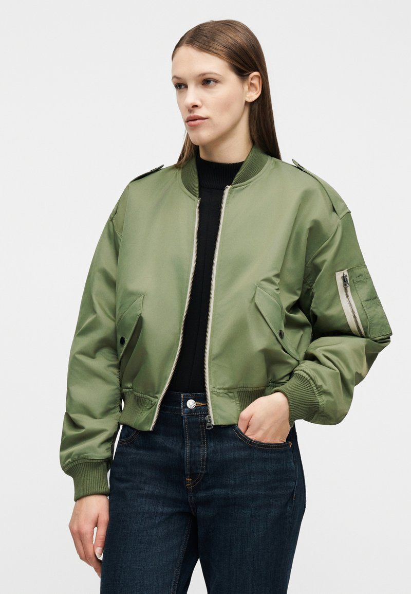 Femme aux cheveux bruns lisses portant une veste bomber verte courte par-dessus un haut noir et un jean bleu foncé, une main dans la poche, regardant vers la gauche.