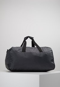 Musta nailonista urheilukassi, jossa on kaksinkertaiset kahvat, suorakulmainen pohja ja vetoketjullinen kansi, asetettuna valkoiselle jalustalle harmaata taustaa vasten.