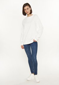 Fehér, oversized pulóver, ejtett vállakkal, kék skinny farmerral és fehér tornacipővel párosítva, egyszínű háttér előtt.