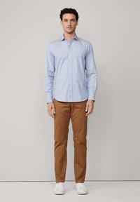 Camisa de botones azul claro con finas rayas verticales, combinada con pantalones marrones y zapatillas blancas. La camisa tiene cuello y mangas largas.