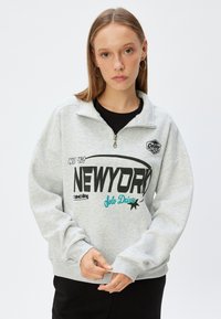 Grå zip-tröja med krage som har en "NEWYORK" grafik i svart och turkos, ribbade muddar och en avslappnad passform. Blandad ulltextur.