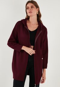 Bordo spalvinė su gobtuvu, pagaminta iš minkštos medžiagos, su užtrauktuku priekyje, ilgomis rankovėmis ir laisvu siluetu, derinama su juodu marškinėliu ir tamsiomis džinsomis.