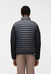 Άνδρας, ο οποίος φαίνεται από πίσω, φοράει γκρι γιλέκο με ραφές πάνω από μαύρο μακρυμάνικο πουκάμισο και καφέ παντελόνι, με φόντο ένα απλό τοίχο.