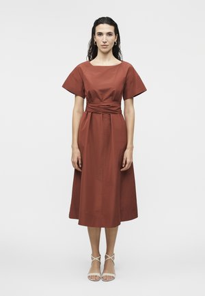 Moteris su tamsiais plaukais dėvi rūdžių spalvos midi suknelę trumpomis rankovėmis ir surauktą juosmenį, derinančią su baltomis striukėmis basutėmis, priešais paprastą foną.