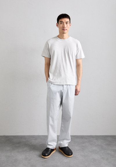 Graues T-Shirt mit kurzen Ärmeln, helle gestreifte Hose und schwarze Holzclogs. Das Modell steht vor einem schlichten grauen Hintergrund.