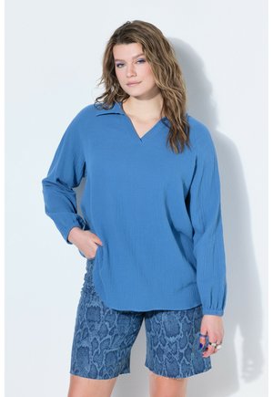 Kobieta z falującymi włosami w luźnej, niebieskiej bluzce z długim rękawem i niebieskich szortach z motywem wężowej skóry, stojąca na tle białej ściany.