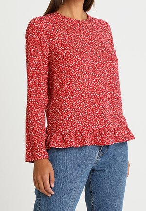 Femme portant un chemisier rouge à manches longues avec imprimé floral blanc et ourlet volanté, associé à un jean bleu en denim.