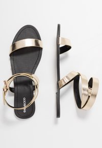 Guldmetalliska platta sandaler med svarta sulor. Har två horisontella remmar och ett cirkulärt ankaremm. Släthet och minimalistisk design.