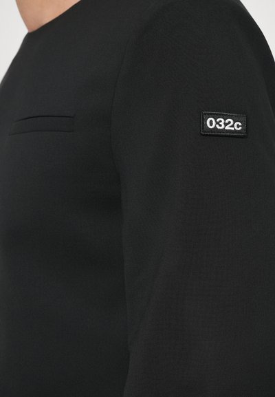 Tricou negru cu mânecă lungă, cu guler rotund, având un mic buzunar pe piept și un patch din material cu "032c" pe mâneca stângă. Textură netedă.