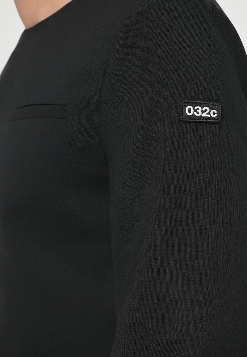 Black long-sleeve top with a crew neck, featuring a small chest pocket and a fabric patch with "032c" on the left sleeve. Smooth texture.