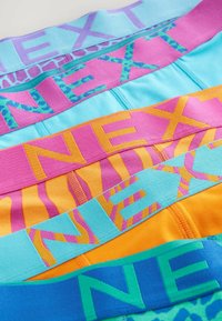 Spalvoti gumeliai su "NEXT" logotipu įvairiais dizainais: šviesiai mėlyna, rožinė su leopardų raštu, oranžinė ir žalia su raštuotais akcentais.
