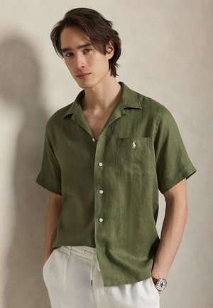 Joven con cabello oscuro viste una camisa de manga corta color verde oliva con botones y pantalones blancos, está de pie frente a un fondo beige liso.