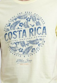 T-shirt en coton jaune pâle avec un graphique circulaire bleu portant le texte "COSTA RICA" et un motif floral. Inclut le nom de la marque "Ritchie Jeans" en bas.