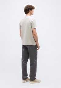 Άνδρας με σγουρά μαλλιά που φοράει μπεζ κοντομάνικο πουκάμισο, γκρι παντελόνι και μπεζ αθλητικά παπούτσια, στέκεται και κοιτά προς τα αριστερά σε λευκό φόντο.