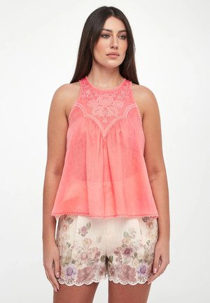 Donna che indossa un top ricamato rosa corallo senza maniche e pantaloncini bianchi con motivo floreale e orlo smerlato, in piedi davanti a uno sfondo bianco semplice.