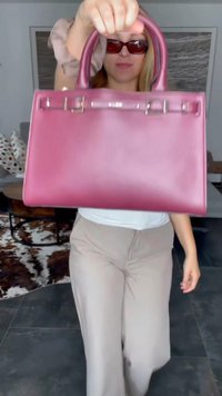 Woman wearing sunglasses holds a large pink handbag toward the camera, standing indoors in beige pants and white top.