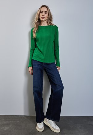Jeune femme avec de longs cheveux blonds portant un pull côtelé vert, un jean large bleu foncé et des baskets blanches, se tenant contre un mur clair.