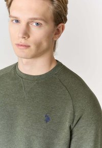 Felpa verde oliva con scollo rotondo e maniche raglan. Presenta un piccolo logo blu sul petto. Realizzata in tessuto morbido e testurizzato.