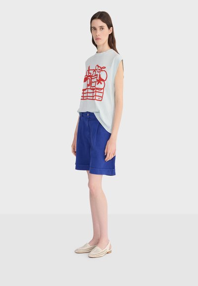 Mujer joven con camiseta sin mangas blanca con diseño abstracto rojo, pantalones cortos azules hasta la rodilla y zapatos beige tejidos sin cordones, de pie de lado.