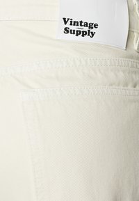 Off-white Denimstoff mit strukturierter Oberfläche. Verfügt über eine sichtbare Tasche und ein Etikett, das "Vintage Supply" in Schwarz zeigt.