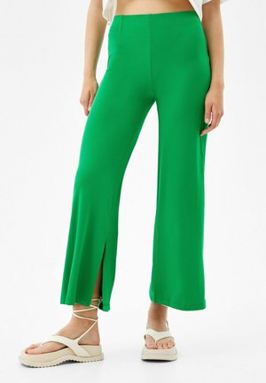 Frau trägt hellgrüne, weit geschnittene Hosen mit vorderen Schlitzen, kombiniert mit beigen, mit Riemchen versehenen Plateau-Sandalen.