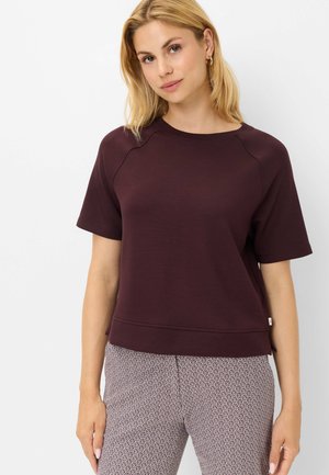 Femme blonde portant un haut violet foncé à manches courtes et un pantalon clair à motifs, debout devant un fond blanc uni.