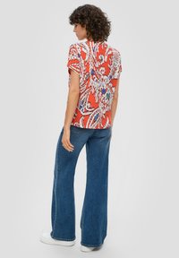 Rotes gemustertes Hemd mit kurzen Ärmeln, florales Design in Blau und Weiß, kombiniert mit hellblauen Schlagjeans und weißen Turnschuhen.