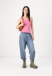 Top sin mangas rosa, pantalones de mezclilla azules holgados con cintura elástica y botas de goma verdes con cremalleras en los tobillos. Bolsa de hombro marrón.