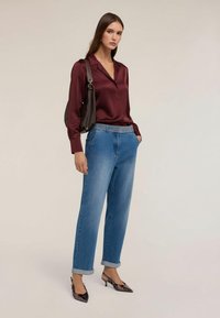 Kobieta w bordowej satynowej bluzce, luźnych niebieskich dżinsach z paskiem ozdobionym ćwiekami, czarnych szpilkach z ostrym noskiem i z czarną torebką na ramię.