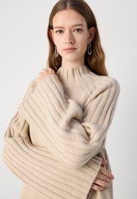 Beige bordás pulóver magas nyakkivágással és széles ujjakra. Puha textúra, látható kötésmintás részletekkel.