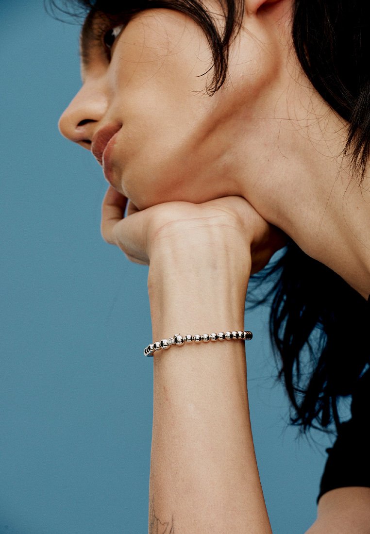 Hõbedane käevõru, millel on ümarate helmedega disain, kantud randmel sinise tausta ees. Sile tekstuur ja poleeritud viimistlus.